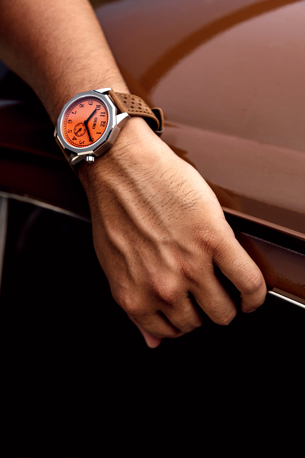 Montre OURO RW-003 associée au bracelet Rallye en cuir café portée au poignet d’un conducteur appuyé sur sa voiture de collection.