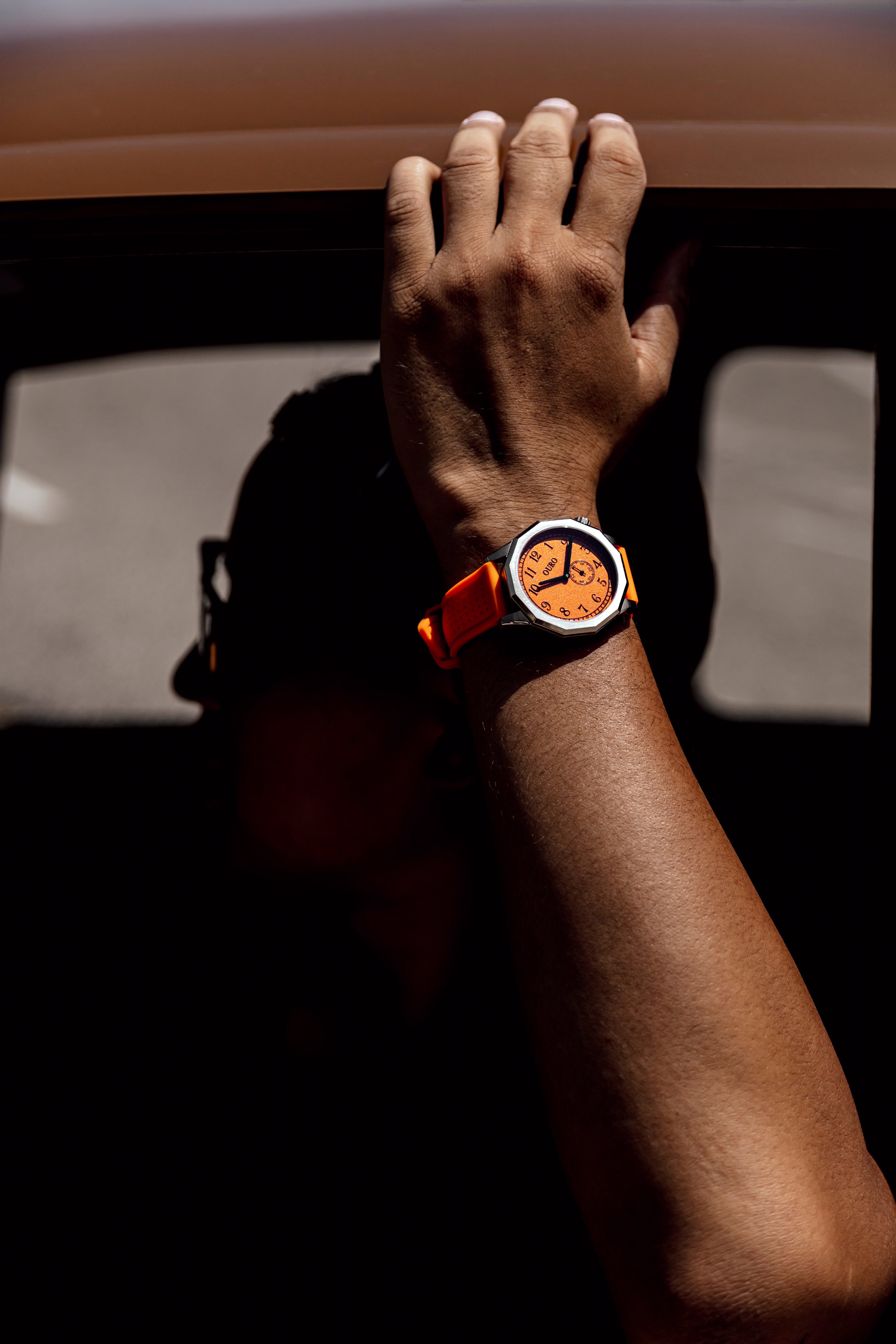 Montre OURO RW-003 associée au bracelet Sport en silicone orange portée au poignet d’un conducteur ayant le bras à la fenêtre de sa voiture de collection.