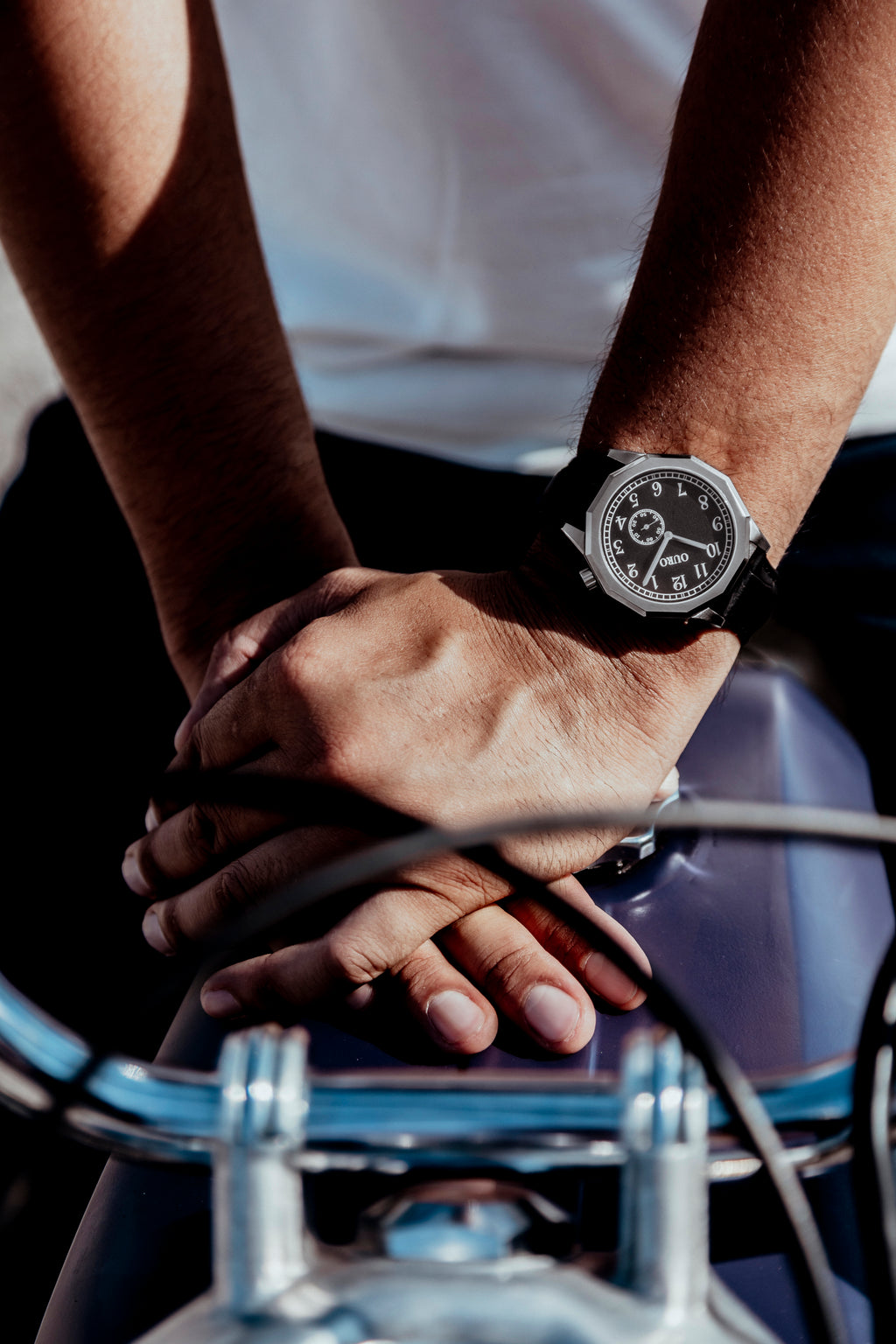 Montre OURO RW-001 avec le bracelet en cuir noir au poignet d’un motard les mains croisées appuyé sur le réservoir de sa moto de collection.