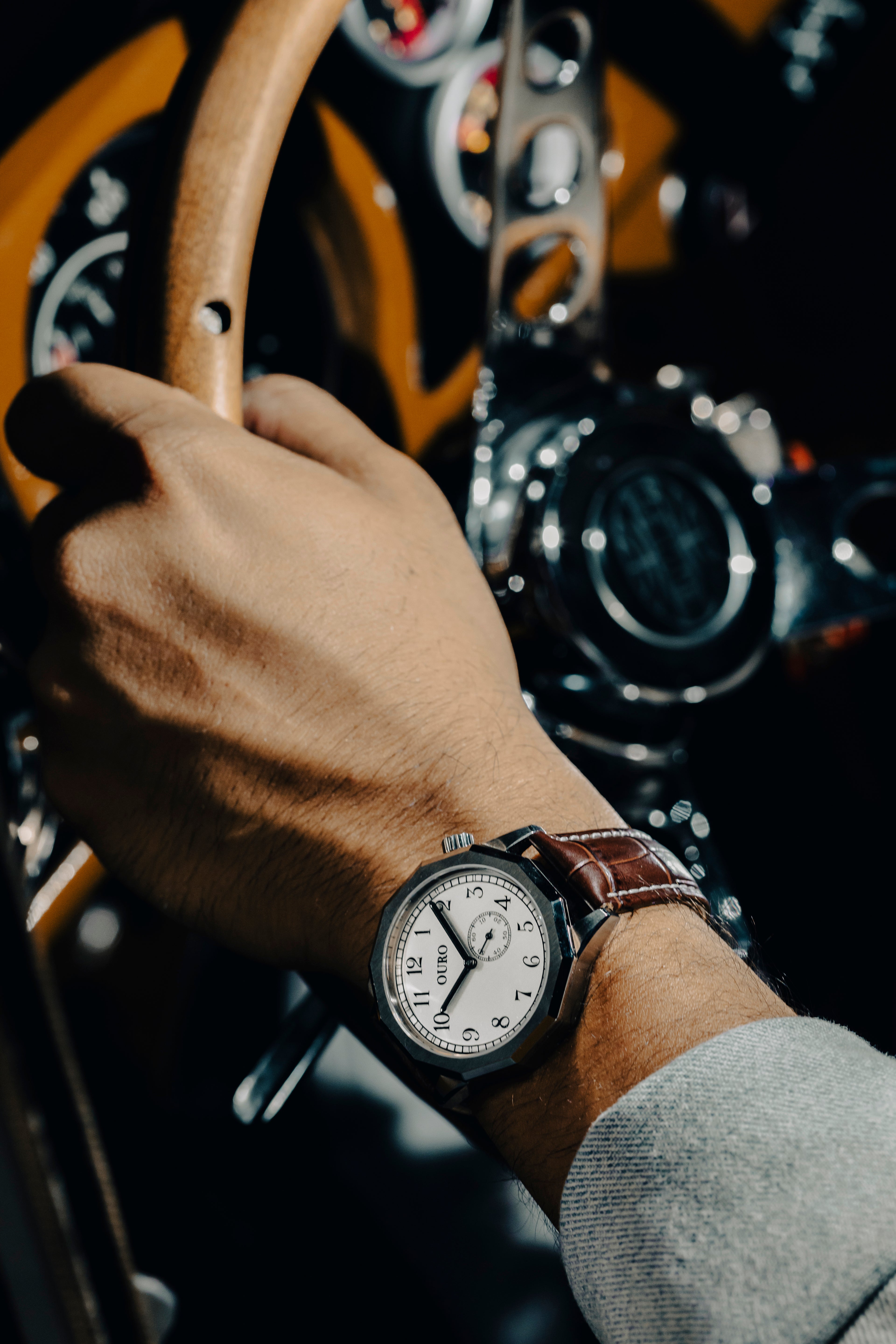 Détail du bracelet Grand Tourisme en cuir marron embossé crocodile monté sur la driver’s watch OURO RW-002 porté par un conducteur tenant le volant en bois de sa voiture de collection.