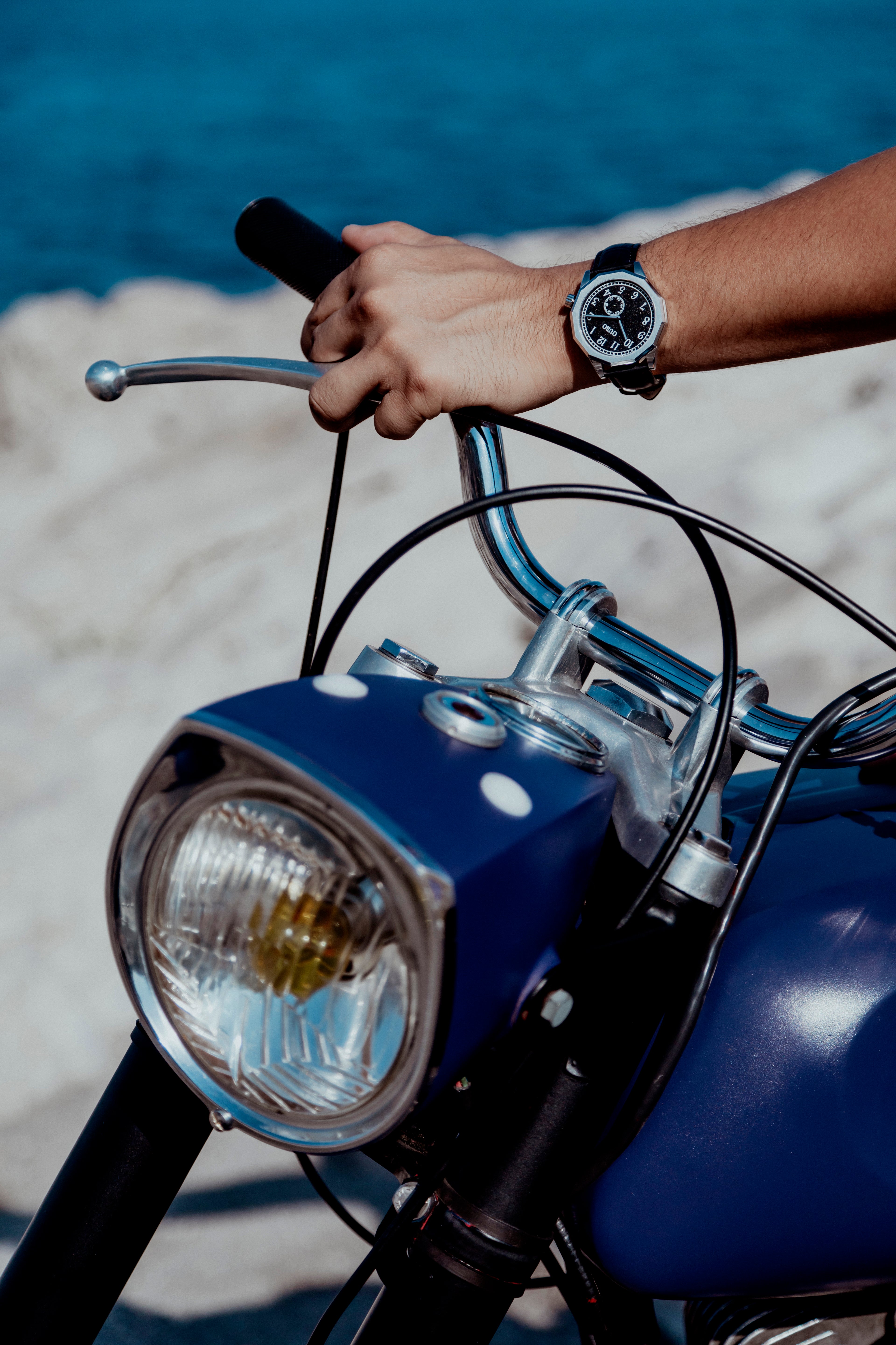 Driver’s watch OURO RW-001 cadran noir décalé à 45° au poignet d’un motard s’appuyant sur le guidon d’une moto ancienne.