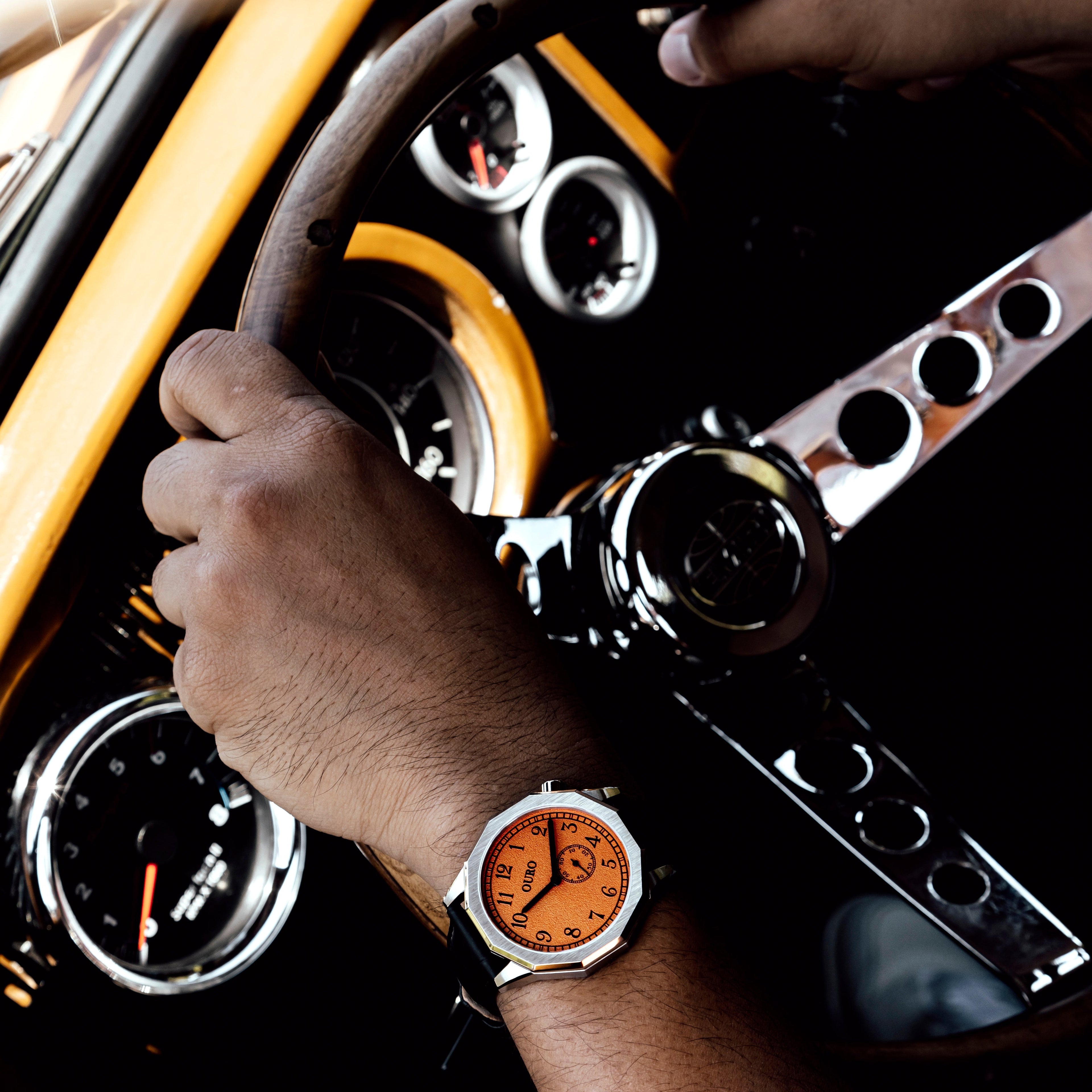Driver’s watch OURO RW-003 cadran orange décalé à 45° au poignet d’un conducteur tenant le volant en bois de sa voiture de collection.